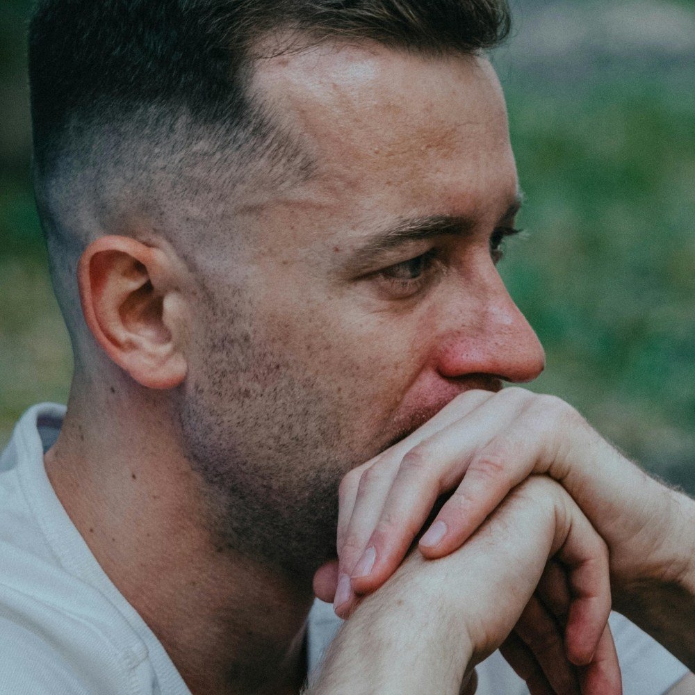 Close-up of a man sitting outdoors with hands clasped near his face, appearing deep in thought.