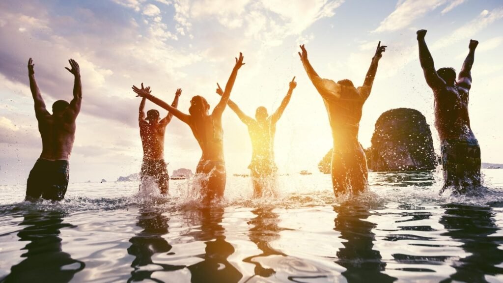 Group of diverse friends celebrating together in ocean water at golden hour with arms raised in joy